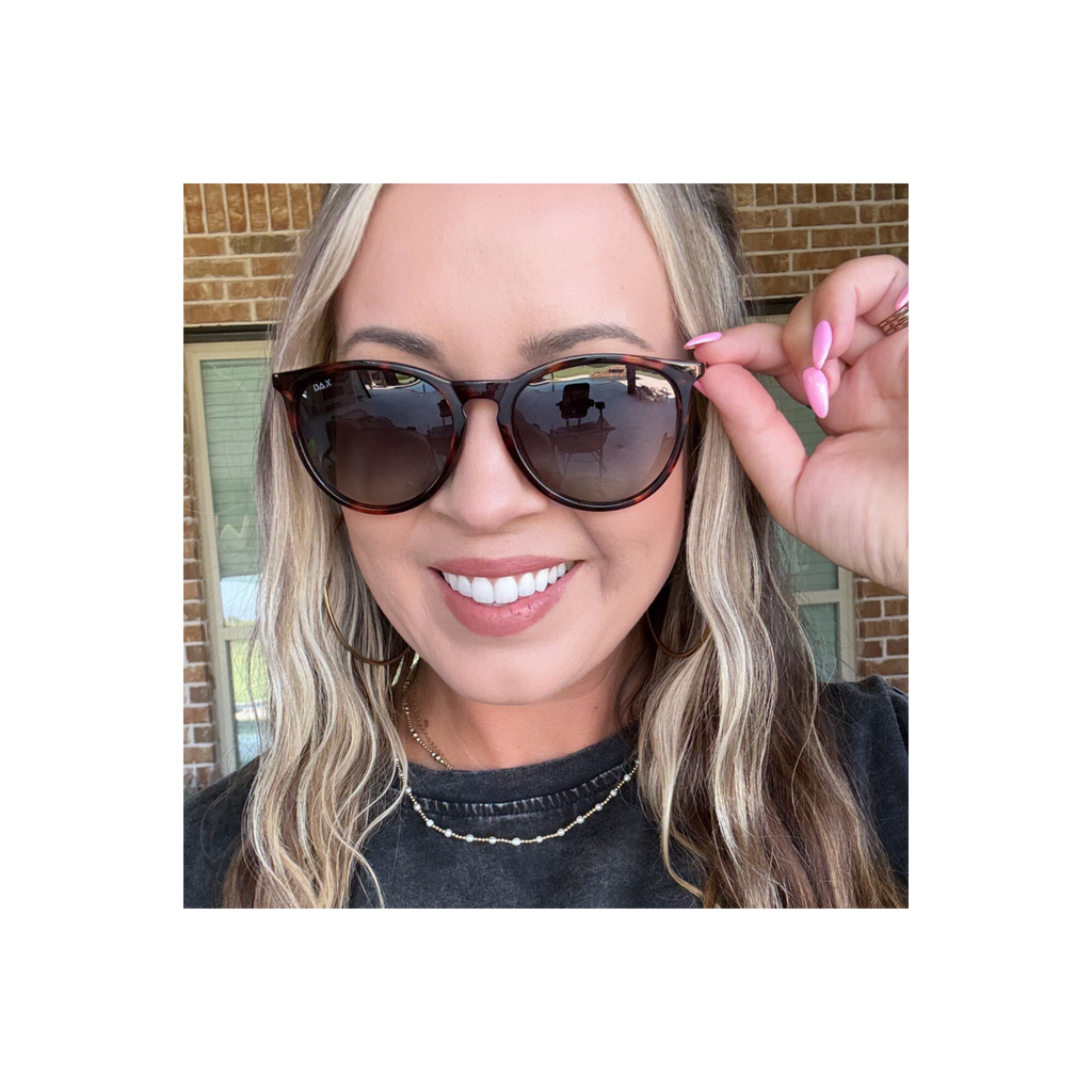 A close-up lifestyle shot of a woman wearing round horn sunglasses with tortoiseshell frames and dark lenses, adjusting the side temple with her hand. She is wearing a black t-shirt and a gold beaded necklace, with a brick wall and window in the background.