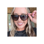 A close-up lifestyle shot of a woman wearing round horn sunglasses with tortoiseshell frames and dark lenses, adjusting the side temple with her hand. She is wearing a black t-shirt and a gold beaded necklace, with a brick wall and window in the background.