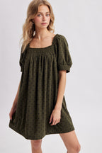 A woman modeling an olive green corduroy puff-sleeve mini dress with navy polka dots against a light gray studio background.