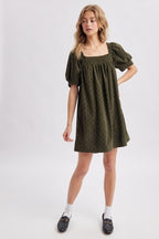 A woman modeling an olive green corduroy puff-sleeve mini dress with navy polka dots against a light gray studio background.