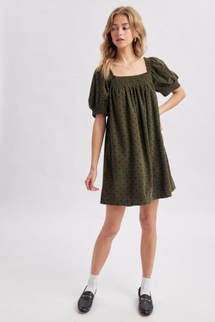 A woman modeling an olive green corduroy puff-sleeve mini dress with navy polka dots against a light gray studio background.