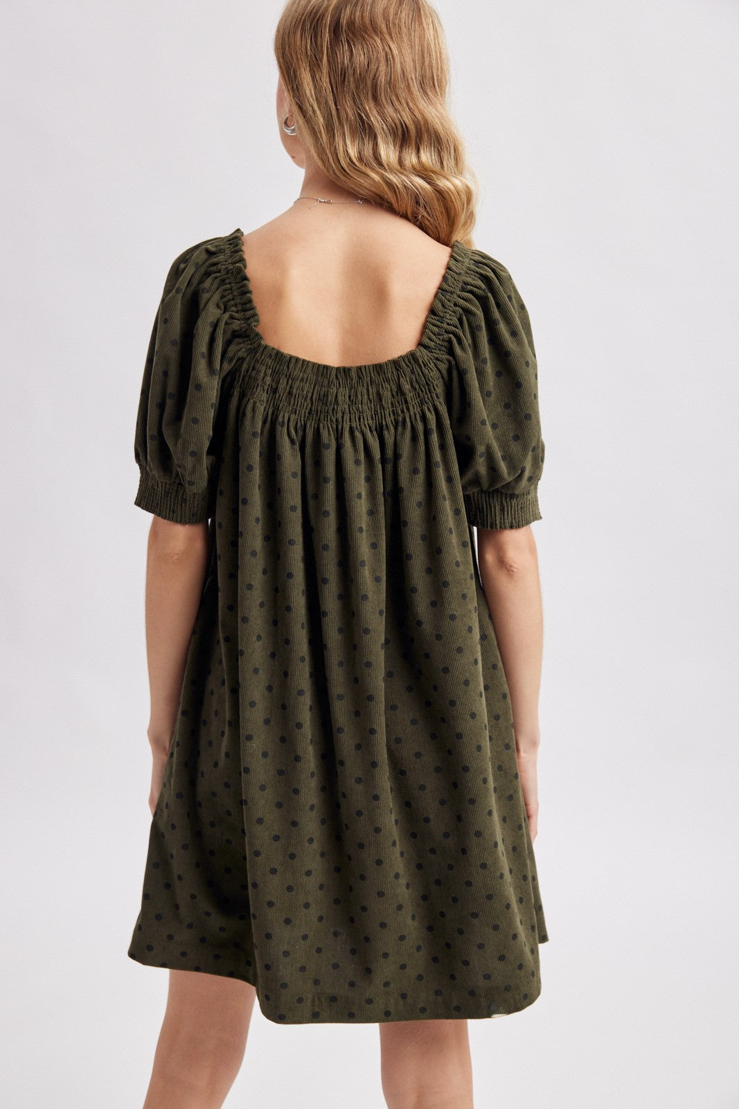 Back view of a woman modeling an olive green corduroy puff-sleeve mini dress with navy polka dots against a light gray studio background.