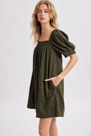 A woman modeling an olive green corduroy puff-sleeve mini dress with navy polka dots against a light gray studio background.