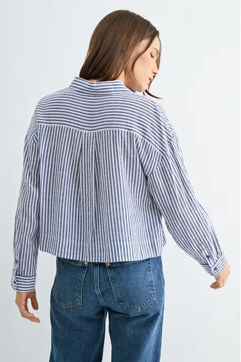 Back view of a woman wearing blue and white textured stripe shirt with front pockets and banded with jeans on a grey background