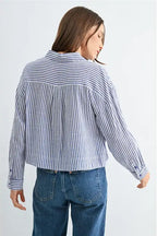 Back view of a woman wearing blue and white textured stripe shirt with front pockets and banded with jeans on a grey background