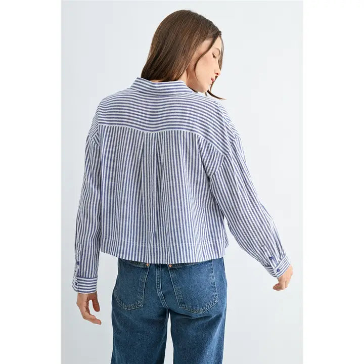 Back view of a woman wearing blue and white textured stripe shirt with front pockets and banded with jeans on a grey background