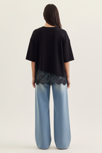 Back view of a woman wearing a black top with asymmetrical lace trim and light blue jeans on a plain background