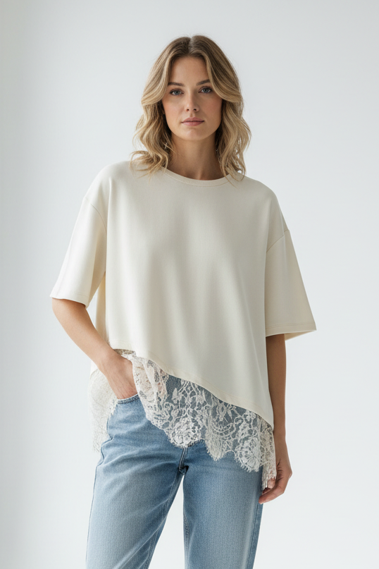 A woman modeling an ecru knit short-sleeve top with a lace hem detail, paired with light blue jeans, against a plain light gray background.