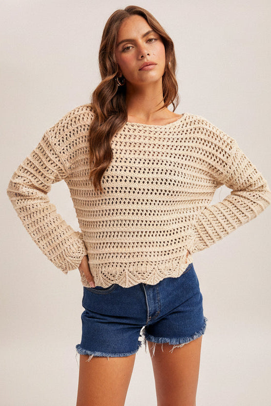 Woman wearing an ecru crochet long sleeve top and denim shorts on a plain background