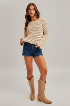 Woman wearing an ecru crochet long sleeve top and denim shorts and boots on a plain background