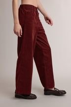 A woman wearing high-waisted, wide-leg trousers in a brick red with a small cream polka dot pattern, styled with dark brown leather loafers against a neutral studio background.