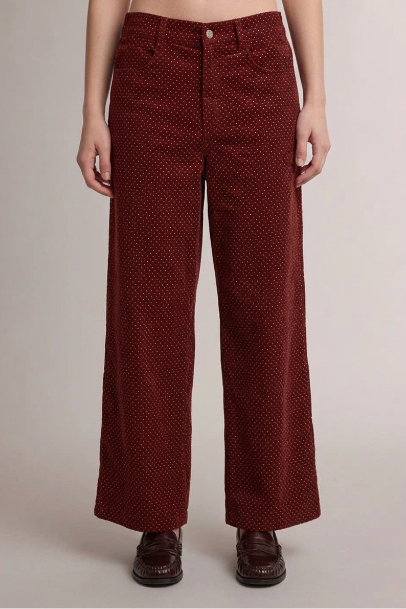 A person wearing high-waisted, wide-leg trousers in a deep burgundy corduroy fabric with a small white polka dot pattern, paired with dark brown leather shoes, against a plain light gray studio background.