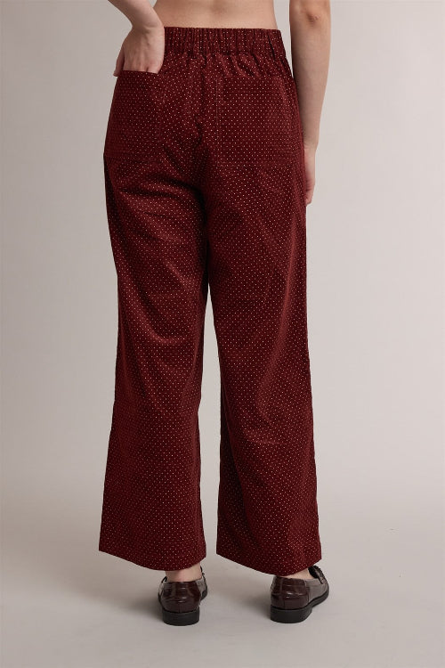 Back view of a woman wearing high-waisted, wide-leg trousers in a brick red with a small cream polka dot pattern, styled with dark brown leather loafers against a neutral studio background.