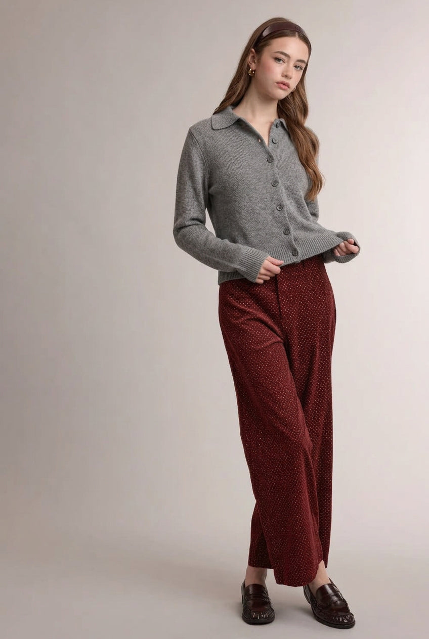 A woman wearing a light gray knit button-up cardigan with a pink argyle pattern, paired with dark brick red mini polka dot small wale corduroy wide-leg pants and dark brown loafers, against a solid light gray studio background.