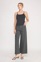 A woman modeling a black cami bralette  paired with dark gray pants against a light gray studio background.