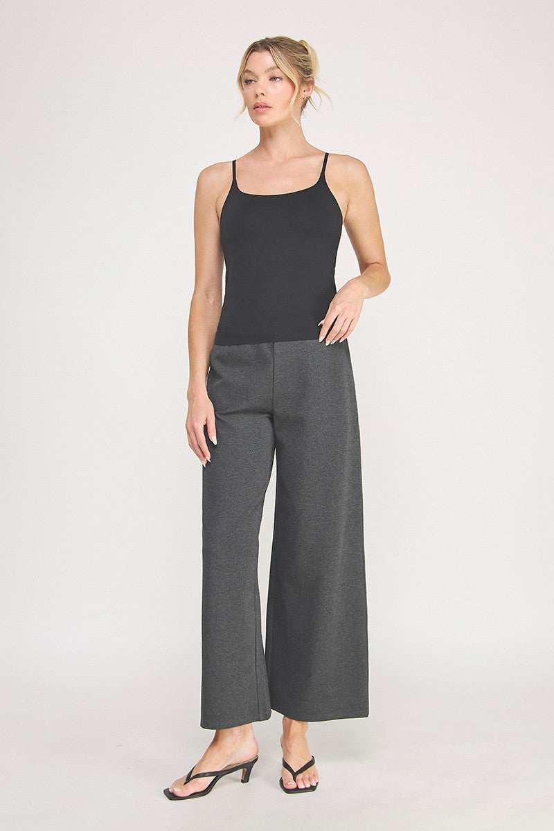 A woman modeling a black cami bralette  paired with dark gray pants against a light gray studio background.