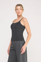 A woman modeling a black cami bralette  paired with dark gray pants against a light gray studio background.