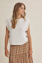 A woman wearing a cream ribbed knit sleeveless vest with brown buttons, paired with a brown and beige plaid skirt and a beige wide-brim hat, set against a solid beige background.