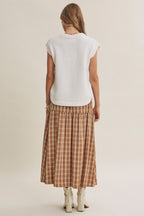 A woman shown from the back wearing a cream sleeveless sweater and a brown and beige plaid midi skirt, paired with white heeled boots, against a plain light beige studio background.