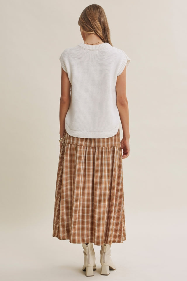 A woman shown from the back wearing a cream sleeveless sweater and a brown and beige plaid midi skirt, paired with white heeled boots, against a plain light beige studio background.