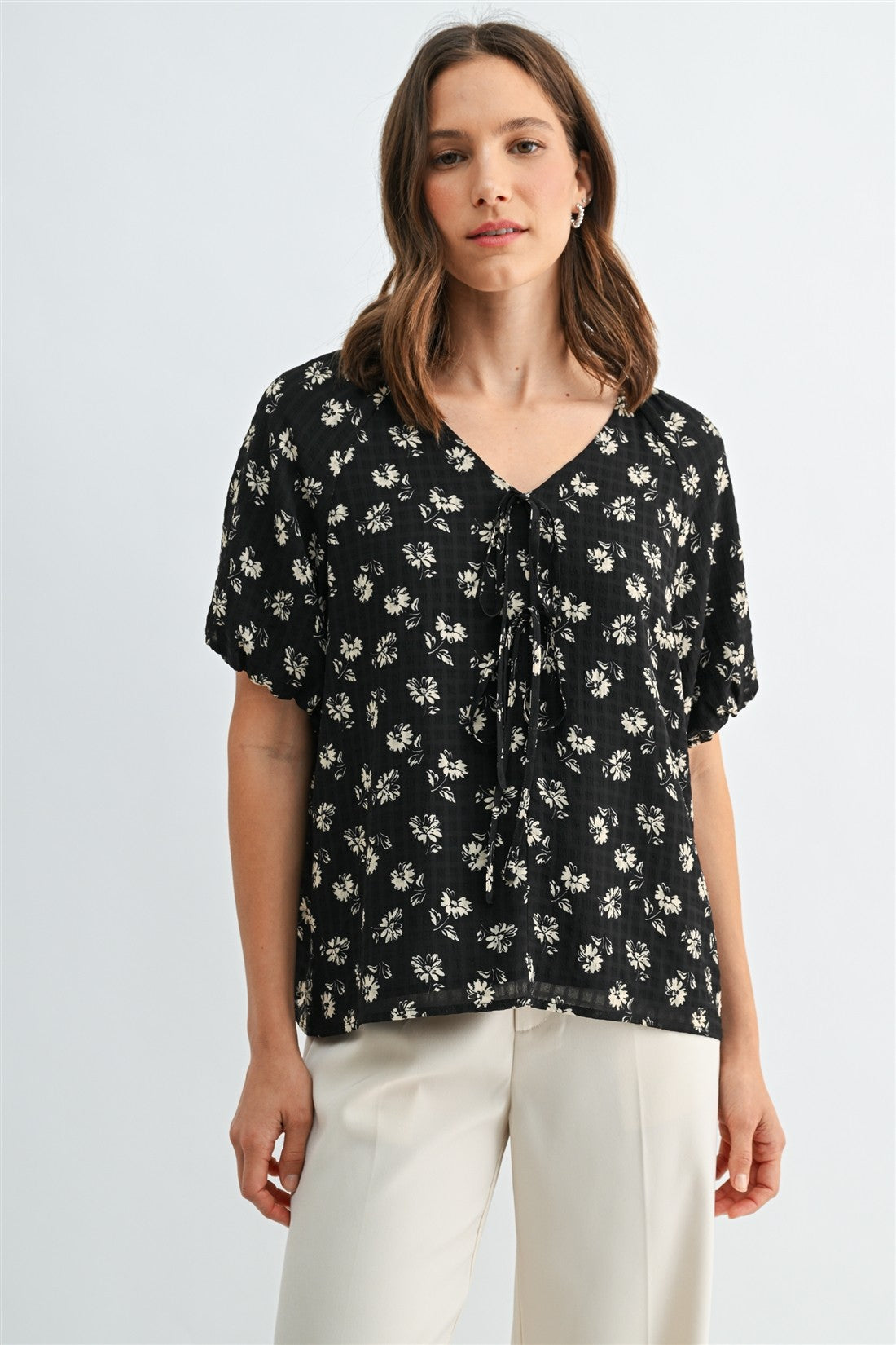 A woman wearing a black and cream floral short-sleeve blouse with a tie-neck detail, paired with cream-colored trousers, against a light gray studio background.