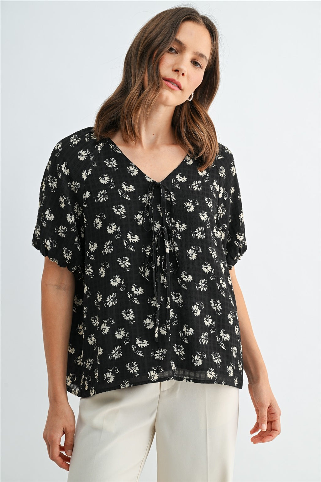 A woman wearing a black and cream floral short-sleeve blouse with a tie-neck detail, paired with cream-colored trousers, against a light gray studio background.