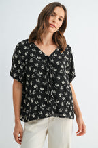 A woman wearing a black and cream floral short-sleeve blouse with a tie-neck detail, paired with cream-colored trousers, against a light gray studio background.