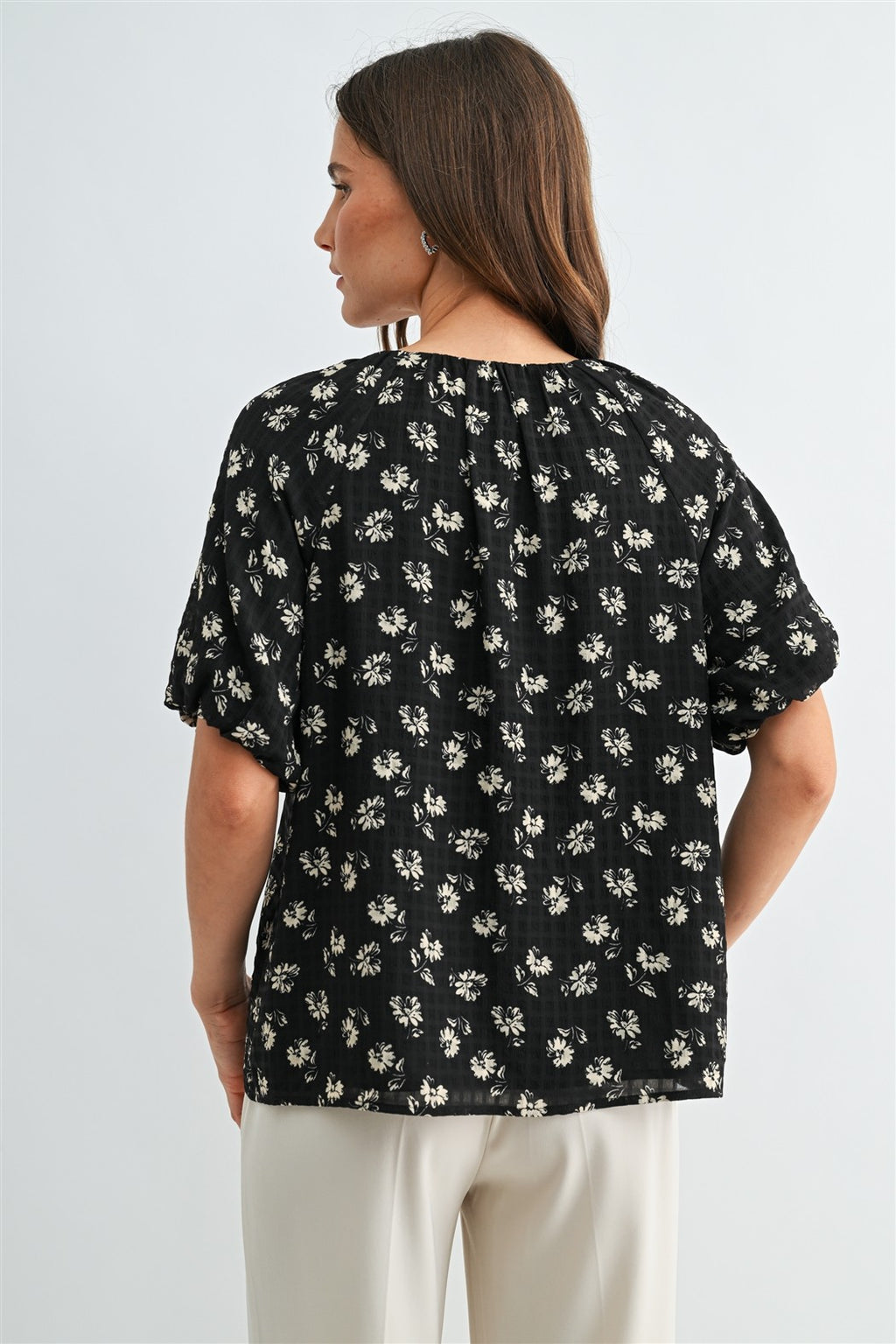 Back view of a woman wearing a black and cream floral short-sleeve blouse with a tie-neck detail, paired with cream-colored trousers, against a light gray studio background.