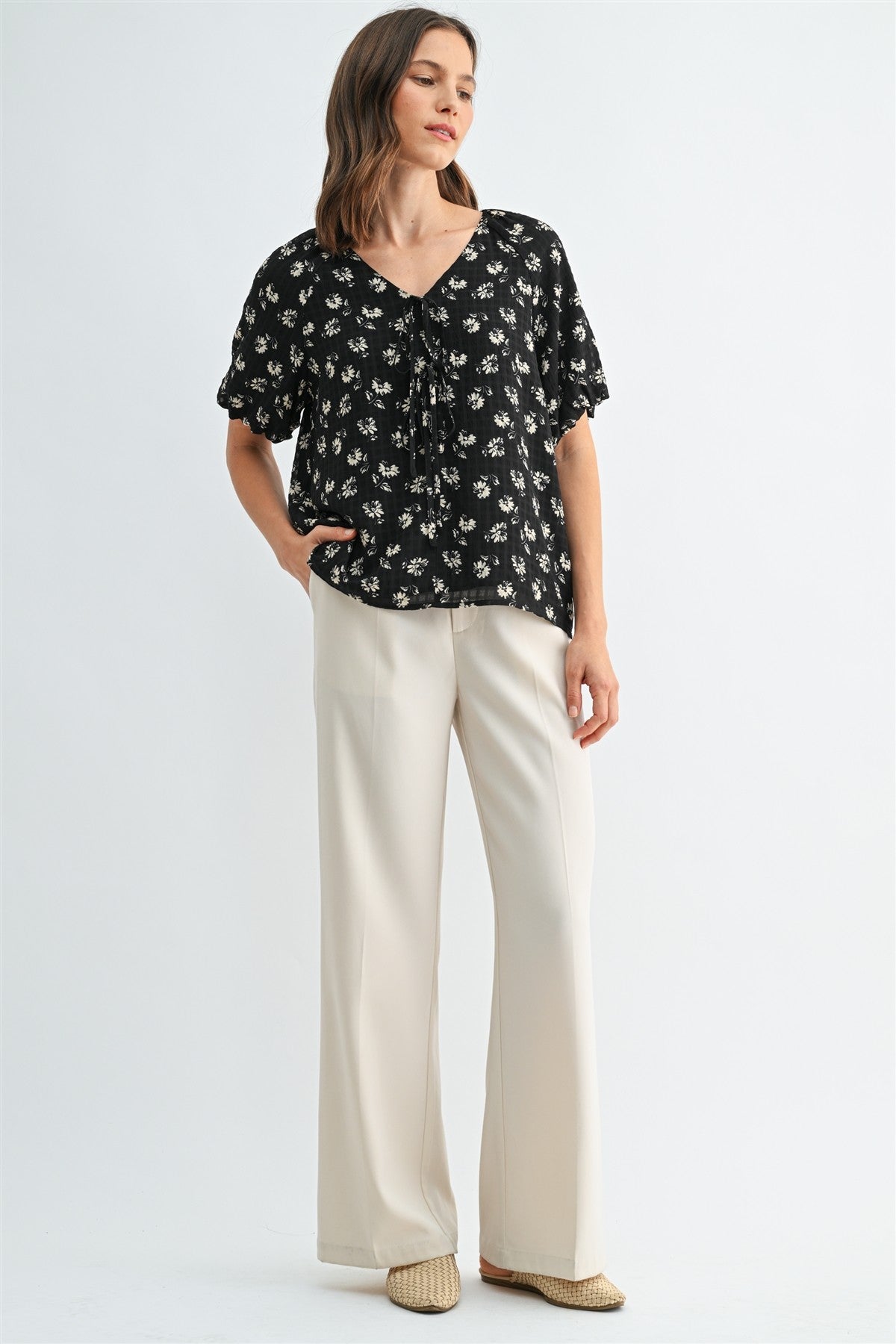 A woman wearing a black and cream floral short-sleeve blouse with a tie-neck detail, paired with cream-colored trousers, against a light gray studio background.