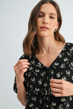A woman wearing a black and cream floral short-sleeve blouse with a tie-neck detail against a light gray studio background.