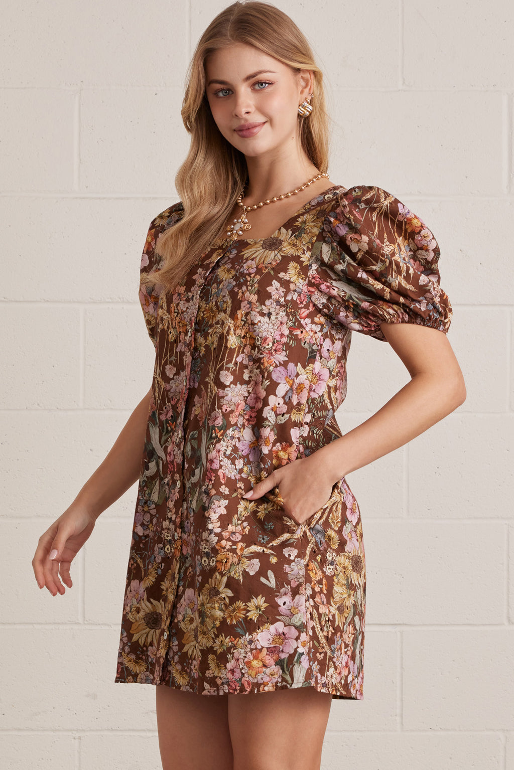 Woman wearing a floral dress with puff sleeves against a white brick wall.