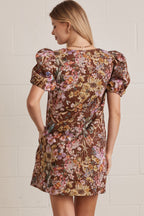Back view of a woman wearing a floral dress with puff sleeves against a white brick wall.