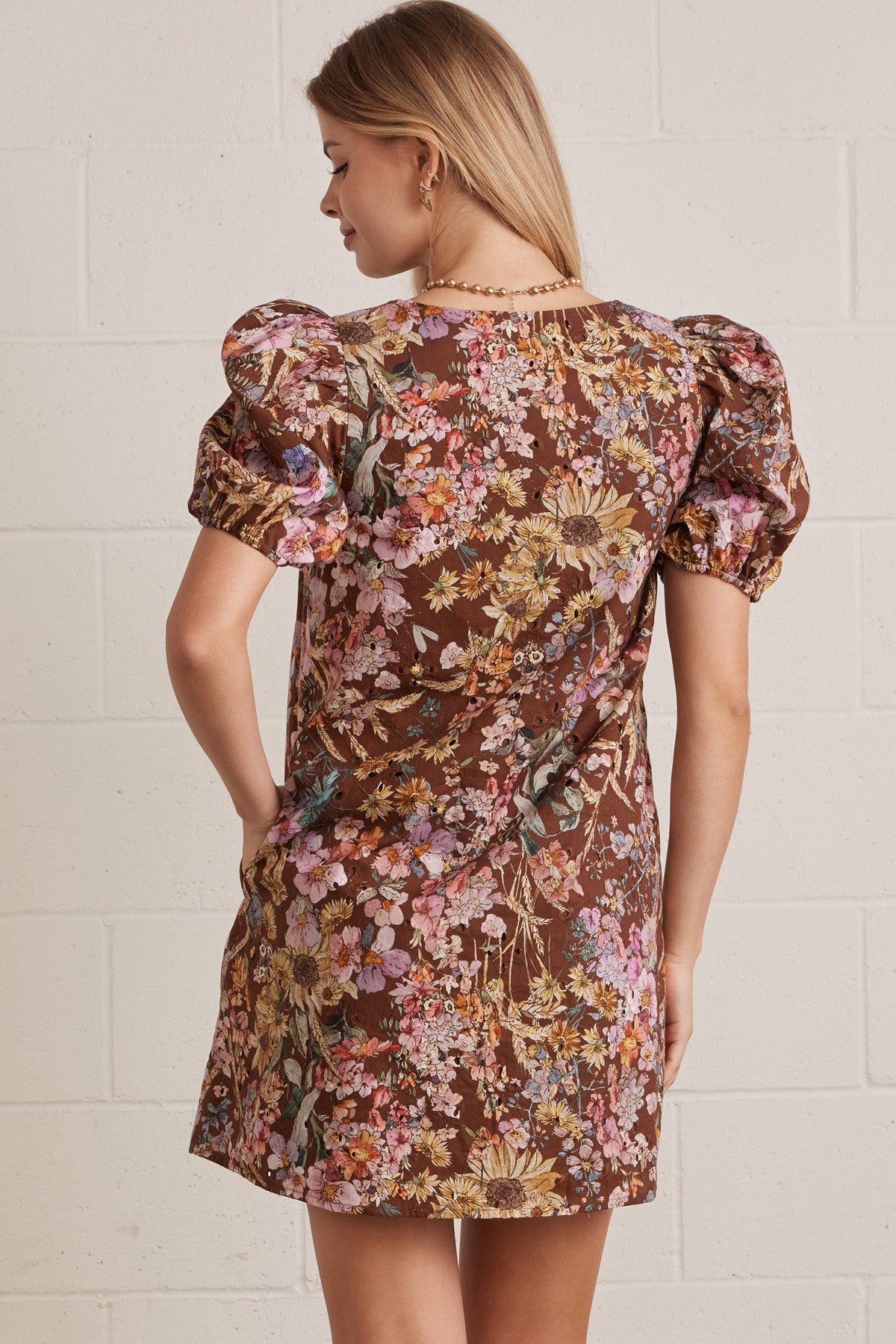 Back view of a woman wearing a floral dress with puff sleeves against a white brick wall.