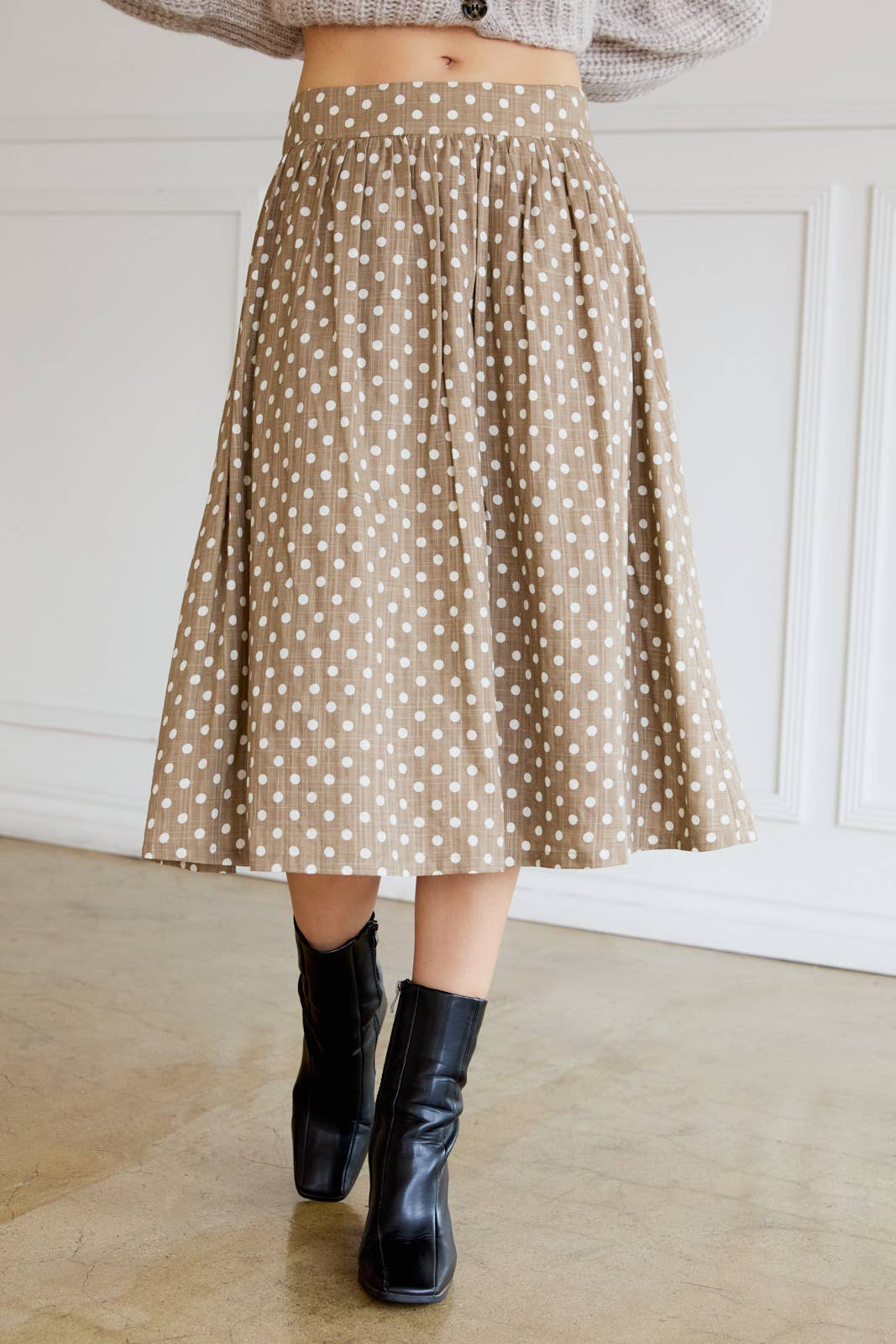 Woman wearing a tan polka dot skirt with cream dots and black boots indoors.