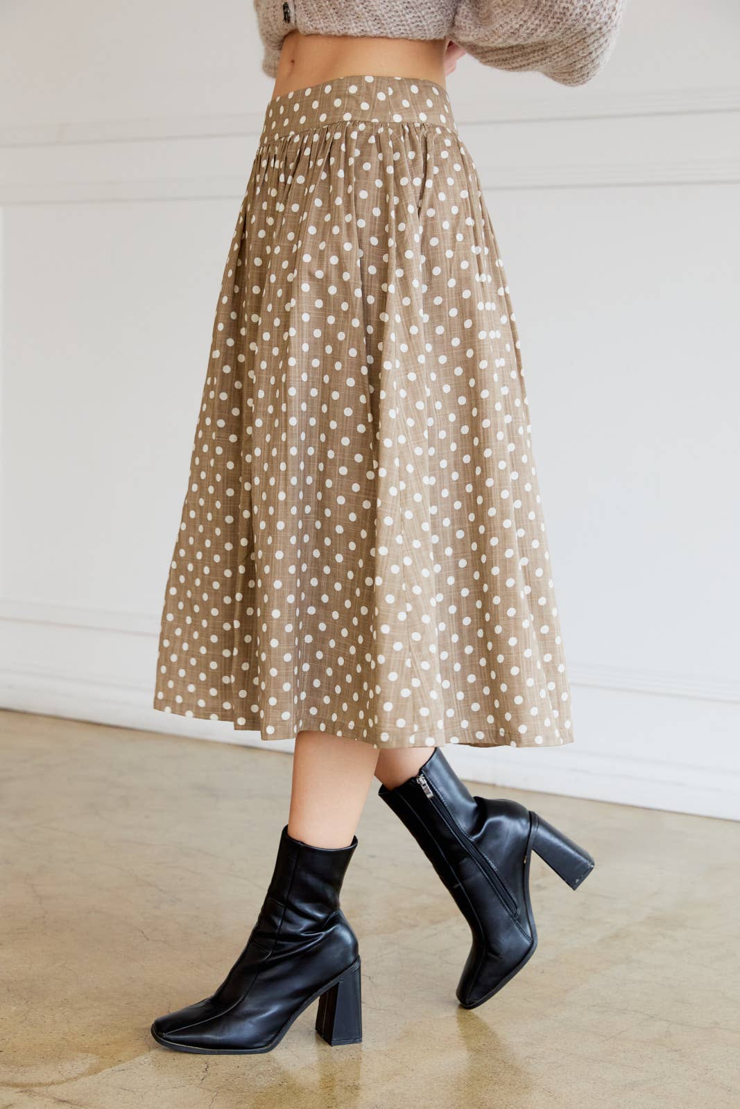 Woman wearing a tan polka dot skirt with cream dots and black ankle boots indoors.