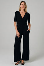 A woman modeling a black short-sleeve jumpsuit with a V-neck, wrap-style bodice, and wide legs, standing against a solid beige studio background.