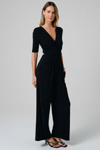 A woman modeling a black short-sleeve jumpsuit with a V-neck, wrap-style bodice, and wide legs, standing against a solid beige studio background.