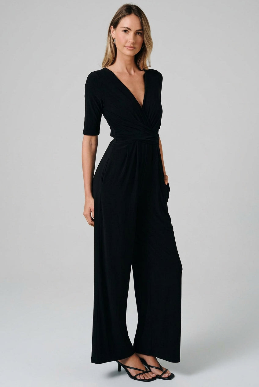 A woman modeling a black short-sleeve jumpsuit with a V-neck, wrap-style bodice, and wide legs, standing against a solid beige studio background.