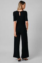 A woman modeling a black short-sleeve jumpsuit from the back, featuring a keyhole cutout at the neckline and wide-leg pants, set against a solid beige studio background.