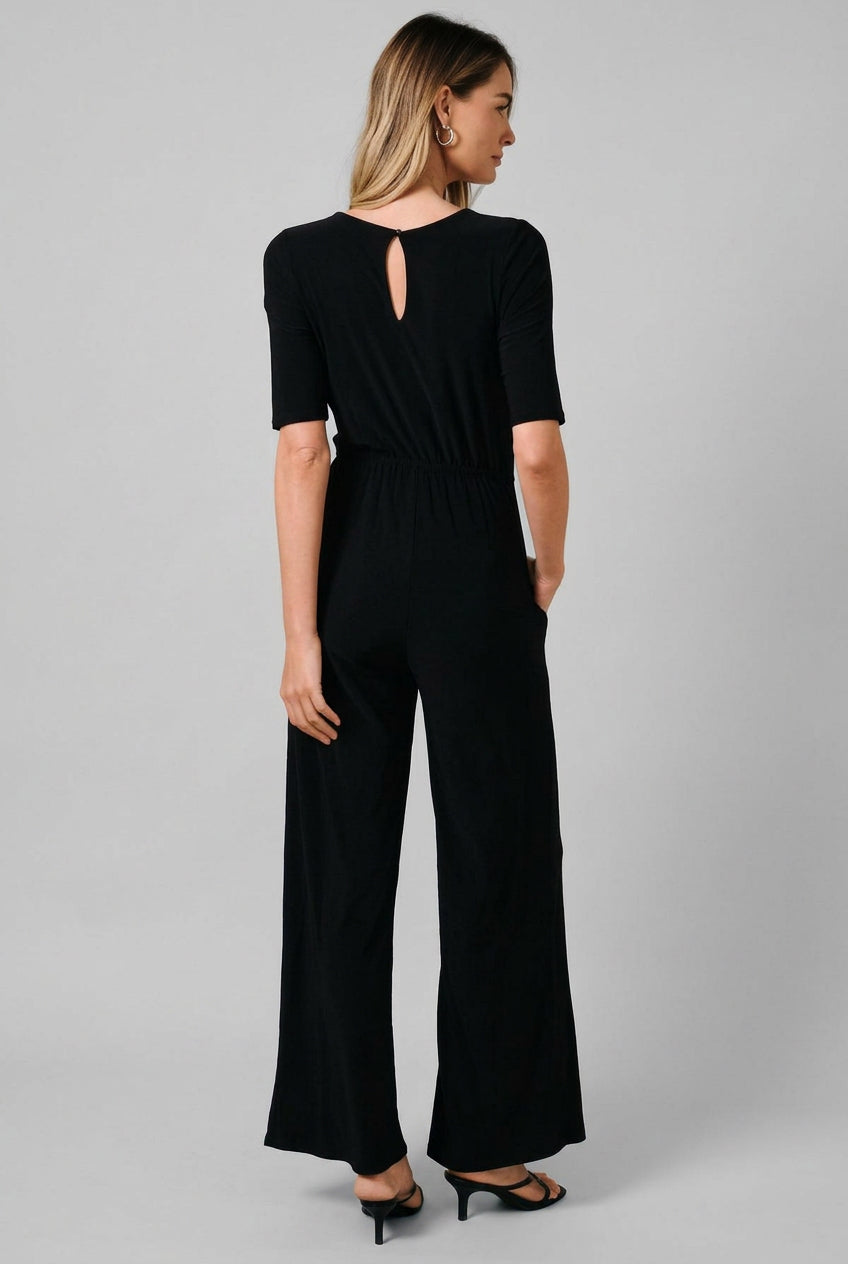 A woman modeling a black short-sleeve jumpsuit from the back, featuring a keyhole cutout at the neckline and wide-leg pants, set against a solid beige studio background.