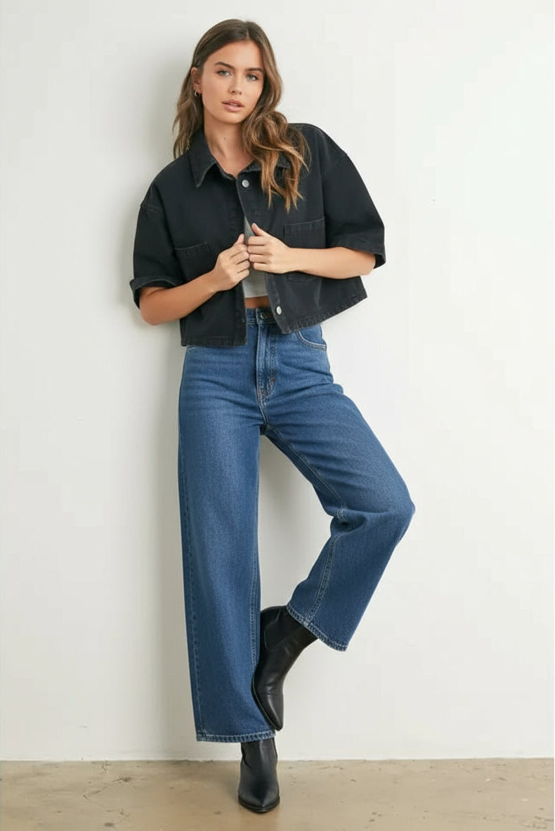 Woman wearing a black denim cropped top, denim, and black boots against a white wall.