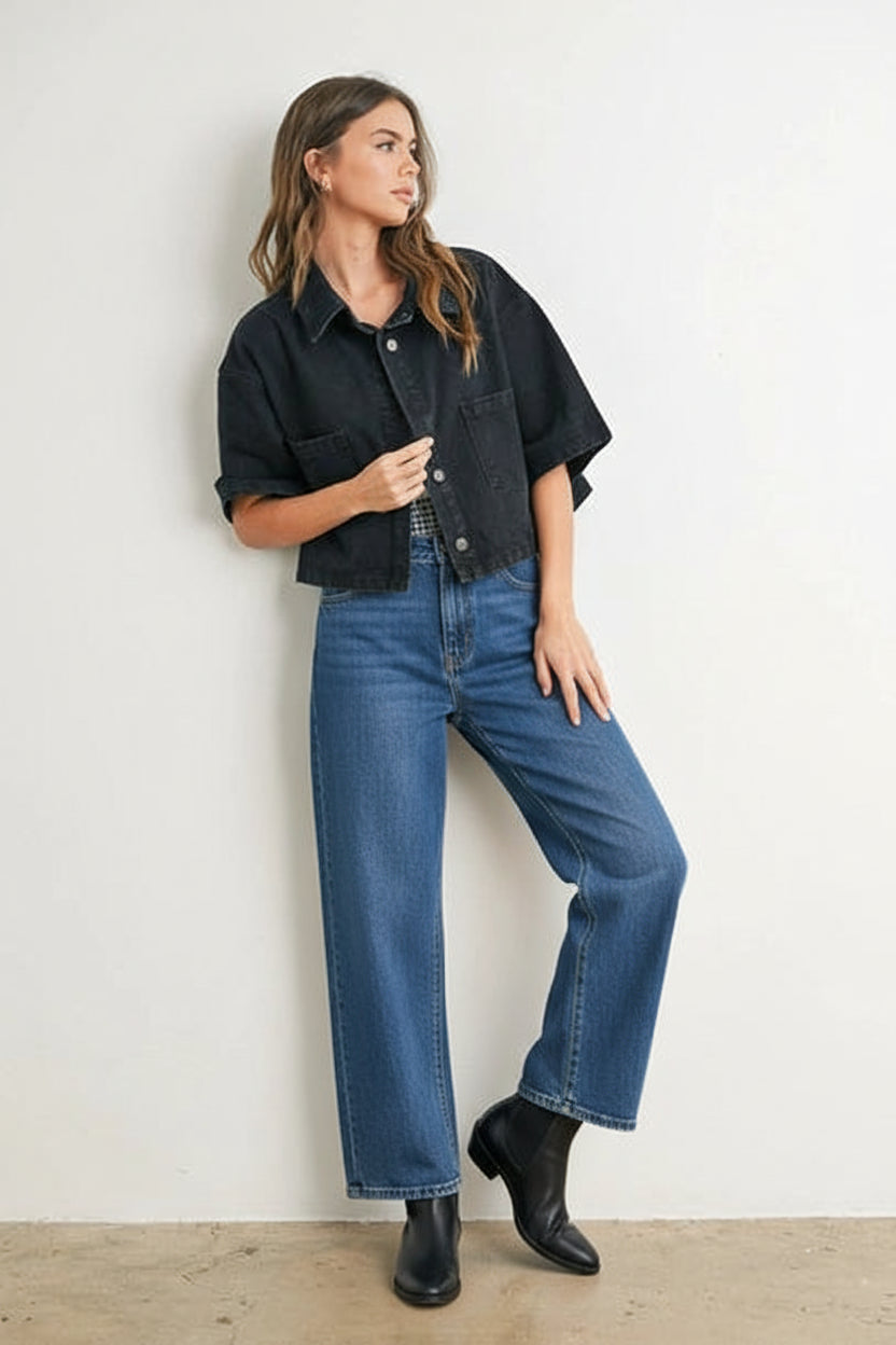 Woman wearing a black denim cropped top, denim, and black bagainst a white wall.ots