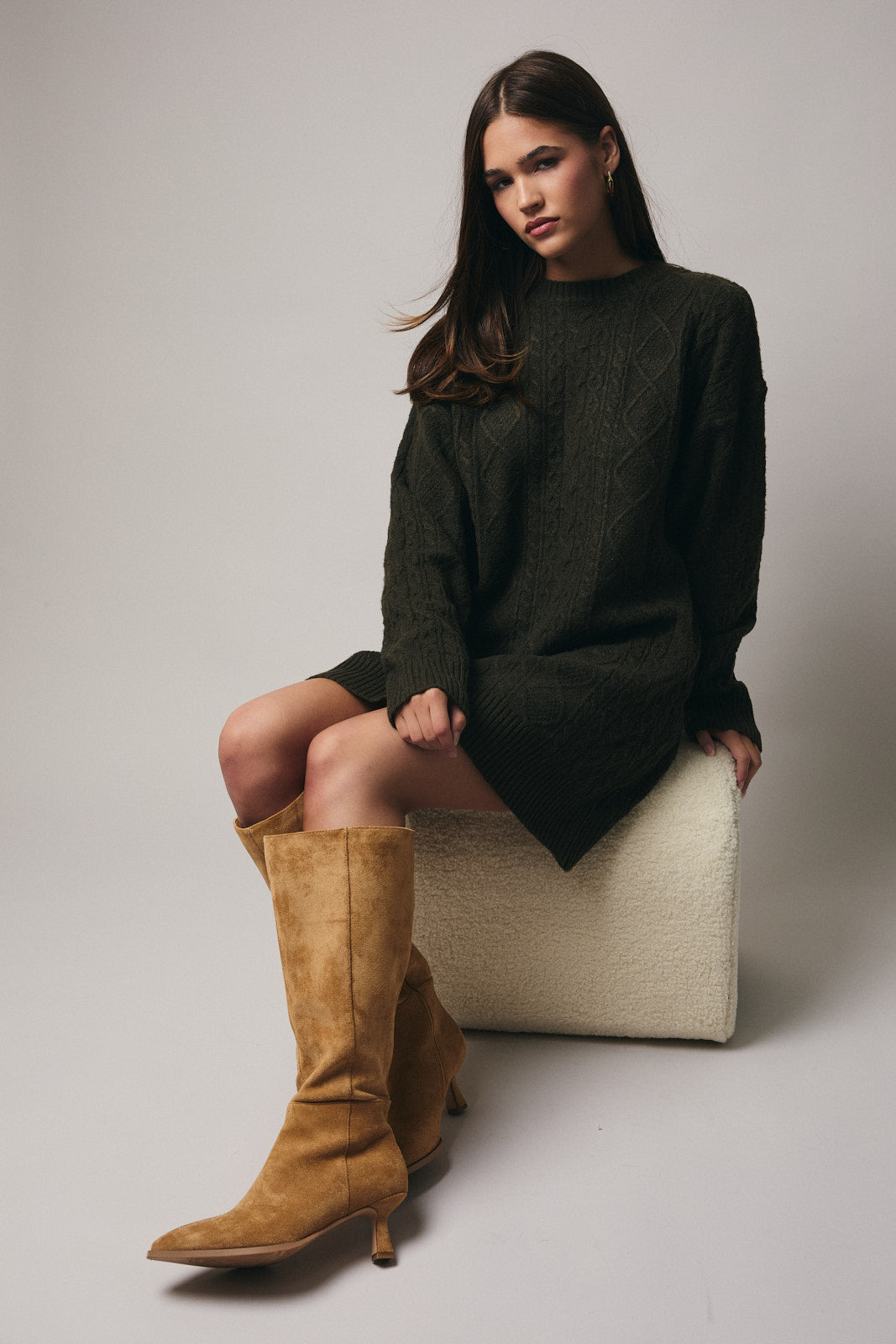 Woman wearing an olive cable knit sweater dress with tan boots against a gray background