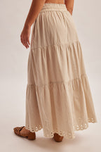Back view of a woman wearing a natural tiered striped skirt with embroidered cutout on a plain background