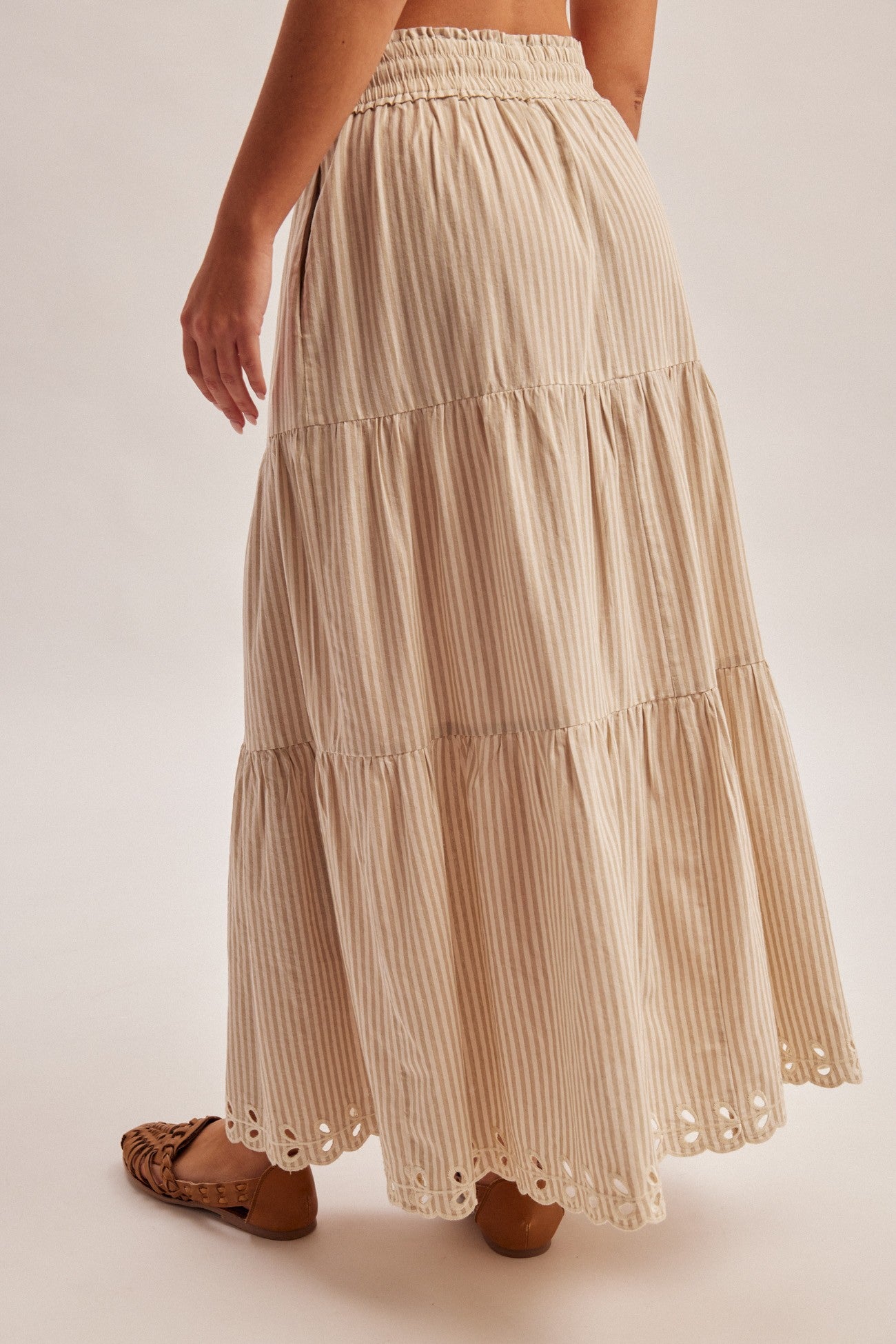 Back view of a woman wearing a natural tiered striped skirt with embroidered cutout on a plain background