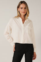 A woman modeling a white long-sleeve button-up shirt with a chest pocket, paired with black pinstriped pants, against a solid beige background.