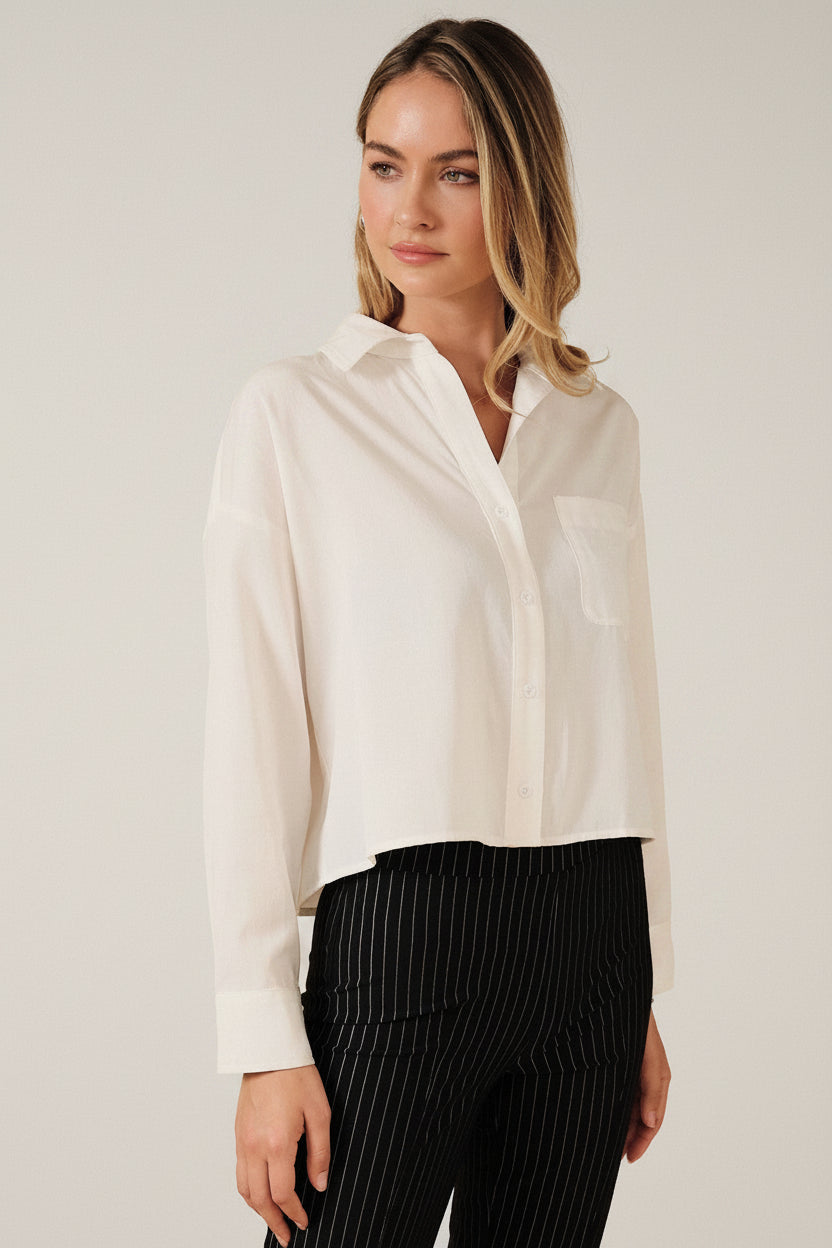 A woman modeling a white long-sleeve button-up shirt with a chest pocket, paired with black pinstriped pants, against a solid beige background.