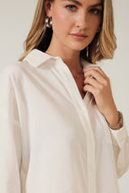 A woman modeling a white long-sleeve button-up shirt against a solid beige background.