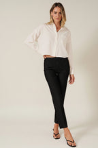 A woman modeling a white long-sleeve button-up shirt with a chest pocket, paired with black pinstriped pants, against a solid beige background.