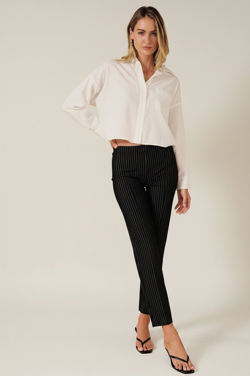 A woman modeling a white long-sleeve button-up shirt with a chest pocket, paired with black pinstriped pants, against a solid beige background.